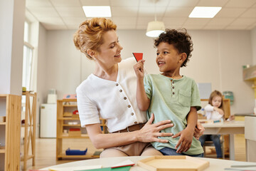 positive african american boy holding triangle near teacher during lesson in montessori school