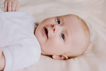 Portrait of happy baby boy in white bodysuit lying on cozy bed at home. Cute little baby smiling and relaxing on bedsheets. Close up top view of caucasian baby newborn in bedroom. Childhood concept