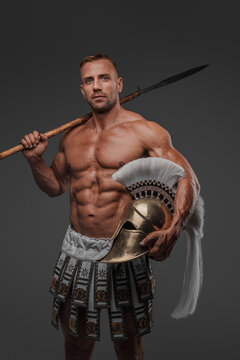 A muscular and attractive male model dressed in ancient pteruges poses with a spear and helmet in hand against a grey background