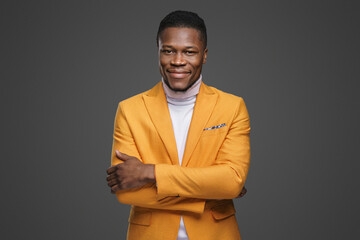 A handsome dark-skinned young man in an elegant yellow blazer and white turtleneck smiling and posing with crossed arms on a grey background