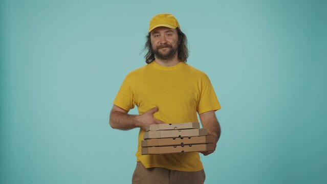 Delivery man in yellow uniform holding a stack of pizza boxes and shows how tasty it is with hungry face expression. Isolated on blue background.