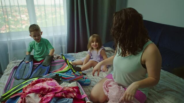 Children Help Mom Put Clothes In A Suitcase For Vacation At Sea. Happy Smiling Family Fooling Around And Having Fun While Packing Their Luggage For A Flight On A Trip. High Quality 4k Footage