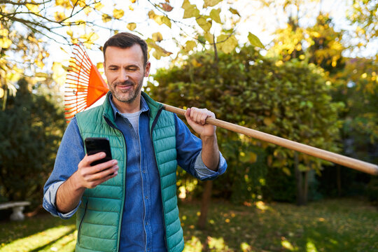 Portrait of adult man standing with leaf rakes in garden using his mobile phone during sunny autumn afternoon