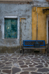 Uno scorcio di Burano, isola della laguna di Venezia, con una panchina blu e una damigiana davanti a una casa disabitata