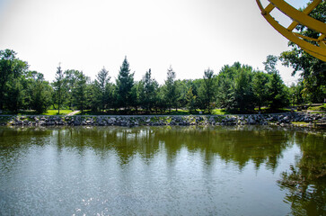 A pond surrounded by a monument and green trees with a lawn © Alexander
