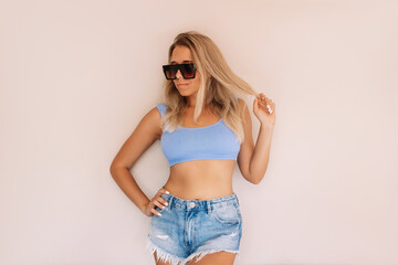 Young caucasian sexy blonde woman in a swimsuit top, denim shorts and sunglasses holding a lock of...
