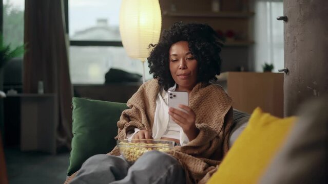 Charming young curly woman with bowl of popcorn hold smartphone browsing scrolling app watching social media feed indoors Happy female texting on her phone enjoying leisure time at cozy home	
