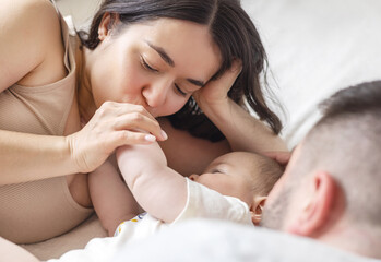 happy young family with baby infant kid boy sitting on bed, cuddling kissing child on hand, cheek, caresses. traditional family lying on bed at home.woman man kid portrait,posing in bedroom