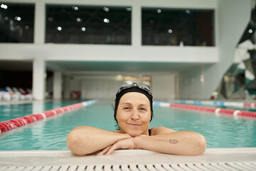 sportive middle aged woman relaxing at poolside, swim cap and goggles, smile, recreation center