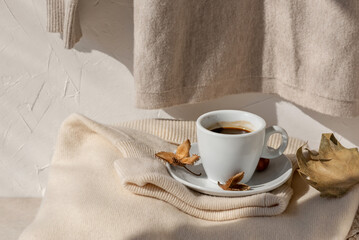 Coffee cup and fall light brown leaves on neutral light beige knitted sweater on beige table and white wall background with sunlight shadow. Aesthetic pastel autumn still life