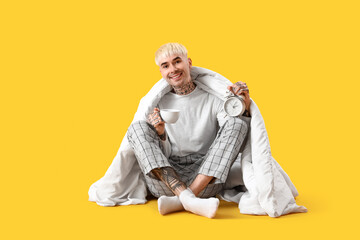 Young tattooed man with blanket, alarm clock and cup of coffee on yellow background