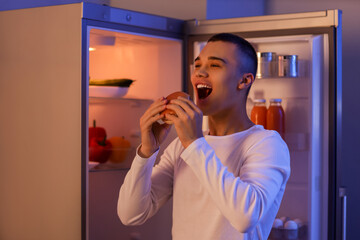 Hungry young man eating burger near open fridge in kitchen at night