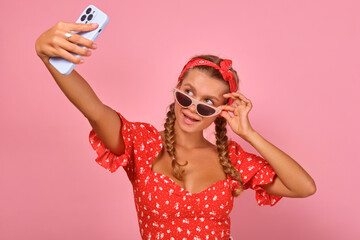 Young pretty tanned Caucasian woman takes selfie on mobile phone and lowers sunglasses from eyes to share photo on social networks stands on pink studio background. Smartphone, apps, internet