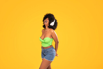Adorable african american woman with white headphones laughing out loud, dancing, yellow vibrant studio background. Real people emotions - obrazy, fototapety, plakaty