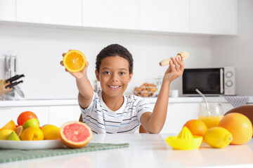 Little African-American boy with juicer and plate of different fresh citruses sitting at table in...