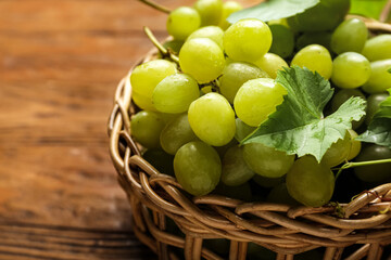 Wicker bowl with sweet green grapes and leaves on wooden background