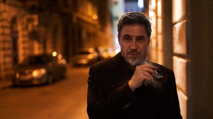 Mid adult man smoking cigar on the street at night wearing black coat. Mature age, middle age, mid adult man in 50s, serious look. Copy space.