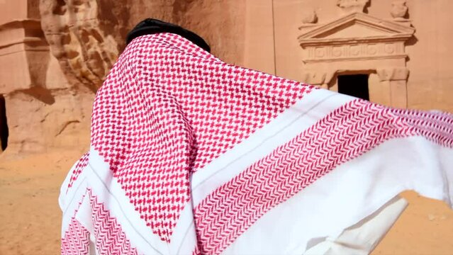 Saudi Arabia arabic man adjusting traditional head scarf in front of a historical landmark in the desert of Al Ula with white clothing and red scarf