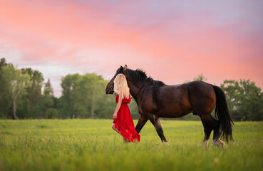 A young woman in a red dress walks with a horse in a field at sunset