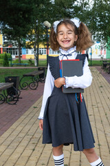 Girl with backpack, school uniform with white bows and stack of books near school. Back to school, happy pupil, heavy textbooks. Education, primary school classes, September 1