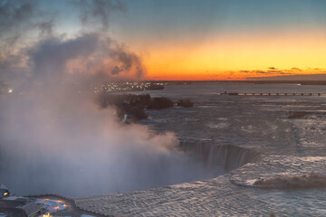 Panoramic view over the Niagara Falls, ON, Canada during winter sunrise with orange sky, water spray from the falls and International Control Dam in distance