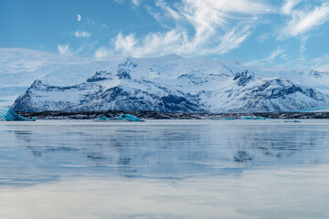 Panoramic view over the mountains surrounding the frozen glacial lagoon of the Jokulsarlon glacier, Iceland with in forefront various blue arctic icebergs and sky with feather clouds and quarter moon