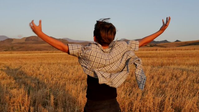 A teenage boy standing in a mowed wheat field at sunset raised his hands up and admires the result. The boy rejoices at the success of his hands raised up. Happy child in a shirt is blown by the wind