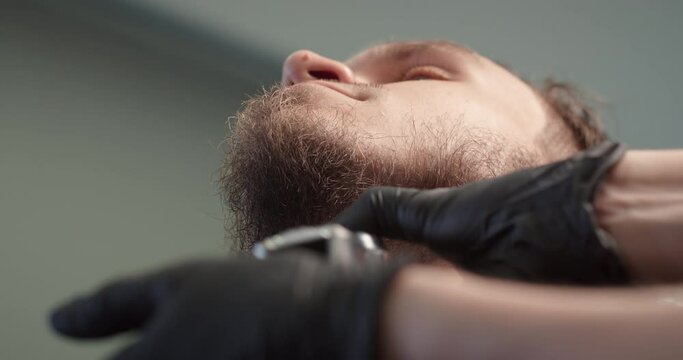 Lower angle shot barber trimming bearded man with shaving machine in barbershop. Hairstyling process. Close-up of a Hairstylist cutting the beard of a bearded male. 4K