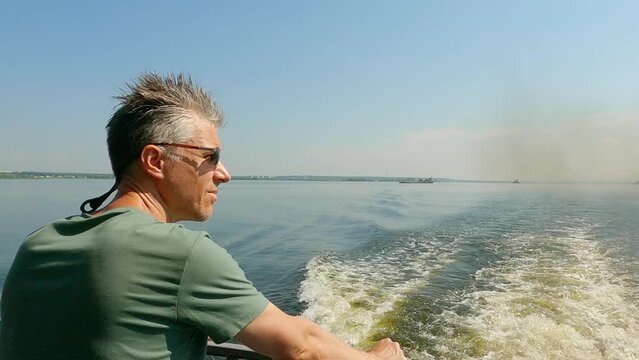 Adult man with a fashionable hairstyle stands at the stern of a river steamer. The steamboat floats along the river on a sunny warm day and smokes. The man is wearing a T-shirt and sunglasses.
