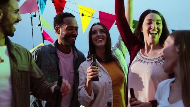 Group of young adult friends having fun dancing on music festival, toasting and drinking alcoholic glasses. Excited and enthusiastic people enjoying their friendship on a rooftop night party. High