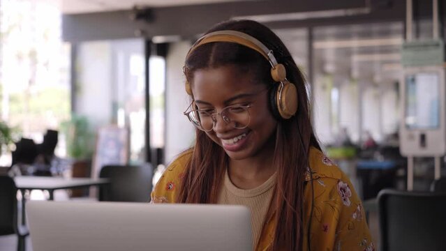 Young African American college student using laptop with headphones. Smiling Latin woman drink hot beverage in campus cafeteria while working or chatting online. People relaxing at class break.