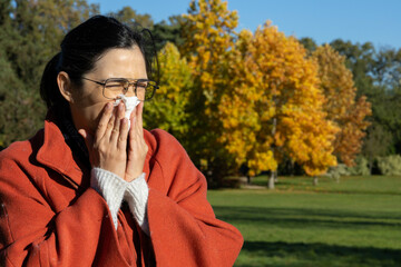 Caucasian brunette middle age woman sneezing and blowing her nose with a tissue outdoors in a park in Paris in autumn. 