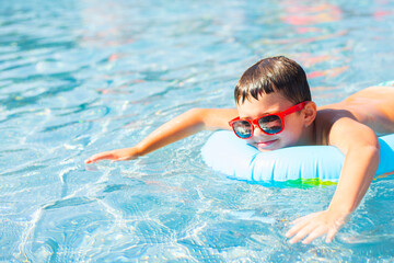 Cute baby boy in red sunglasses swims with a rainbow inflatable ring in a clean pool - 639423075