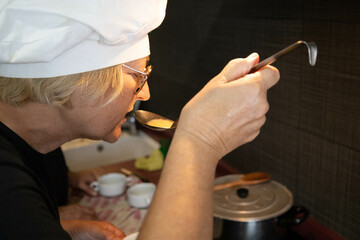 Elderly retired woman in her 60s, blonde and Caucasian, wearing glasses, cooking in her spare time, for her grandchildren. Hungry person tasting the food.