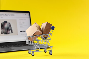 Online store. Mini shopping cart, parcels and laptop on yellow background, selective focus. Space...