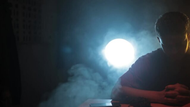Film-like dolly shot of a man in a red shirt sitting on a table preparing for a stealth mission packing his weapons and knives with creative dramatic lighting technique, HD - 24 fps - Prores