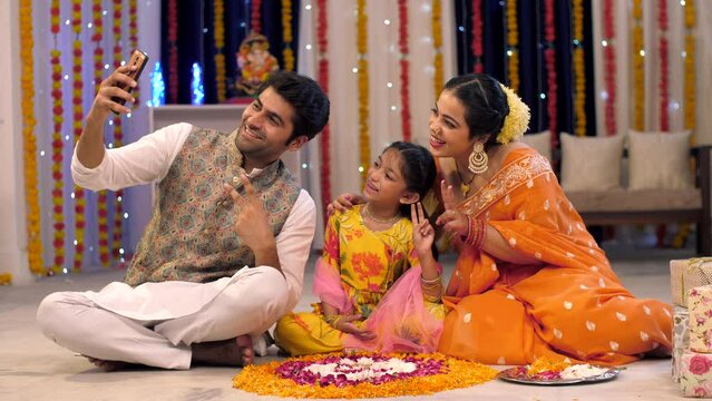 Diwali festival  Indian lovable family taking selfies or self photographs at home -  colorful  decorated background. A family taking photographs at an apartment with rangoli and gifts kept in a liv...