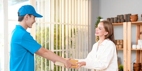 Asian messenger guy in blue uniform with cap giving postal to handcraft business seller lady with smiling face and kind service from fast shipping in small e-commerce office