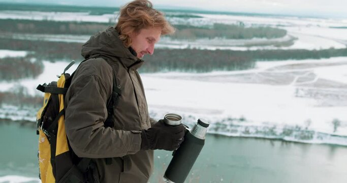 hicker in winter drinks tea in nature, hiking in the mountains in winter, guy pours tea from thermos into cup, lunch in nature in a camp Slow motion. Climbing mountain in winter. Survival in the wild.