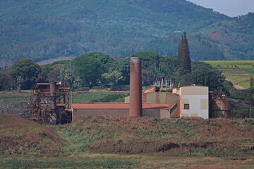 The industrial smokestack and corrugated metal buildings of a former sugar cane processing factory in Hawaii are shown during the day.