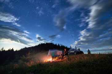 Camping in mountains under evening cloudy sky. Young man having a rest next to campfire, near...
