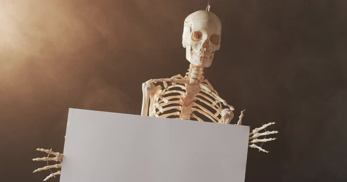 Video of close up of halloween skeleton holding white board with copy space on brown background