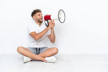 Young blonde man sitting on the floor isolated on white background shouting through a megaphone