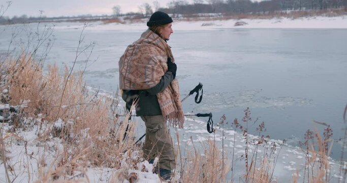 hicker holding Nordic walking poles watching flowing melting ice in the river in spring Slow motion Traveler standing resting on the bank of river adventure, guy feels cold, wrapped on tipped