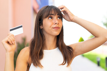 Young woman holding a credit card at outdoors having doubts and with confuse face expression