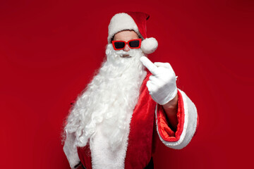 rude santa claus in cap and festive glasses showing middle finger on red background, angry man in...