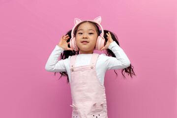 little asian girl in pink children's headphones listens to music and smiles on pink isolated background, korean child