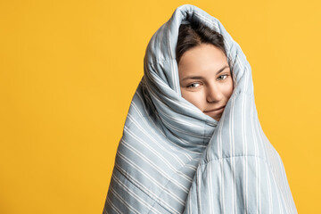 Smiling teenage girl wrapped in warm blanket peeking out with sleepy eyes isolated on studio yellow...
