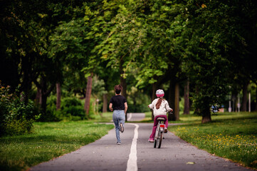 Young mother in jeans and black t-shirt running and playing with her daughter in summer parks while girl riding a bike