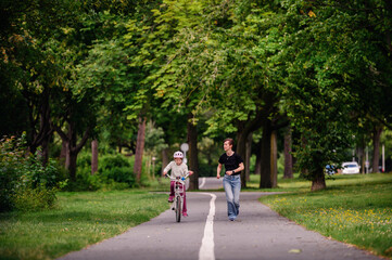 Young mother in jeans and black t-shirt teaching and playing with her daughter in summer parks while girl riding a bike